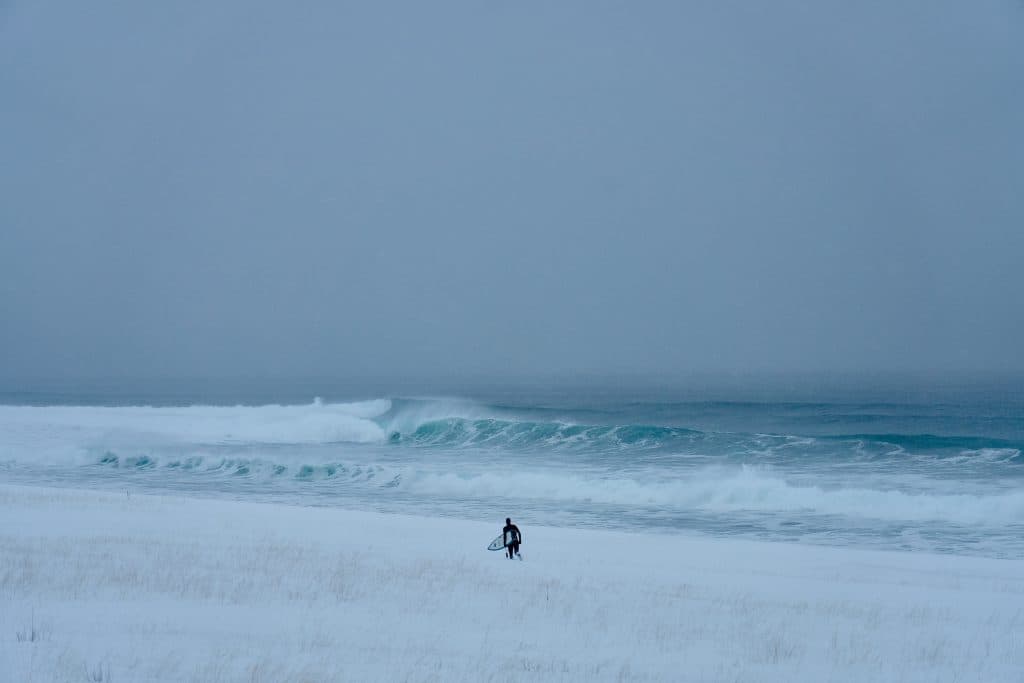 Tim Latte går genom djup snö på väg ut till havet med sin surfingbräda under armen.